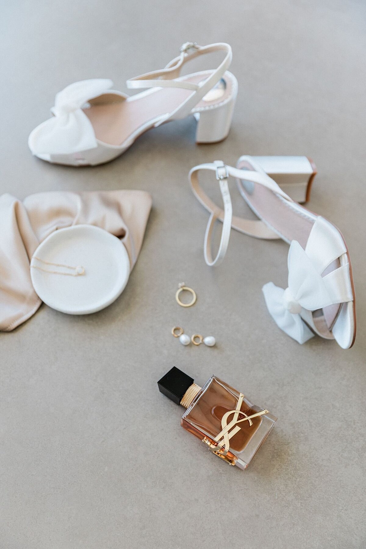 Elegant bridal flatlay featuring white block heels with bows, gold jewelry, pearl earrings, a silk hair ribbon, and YSL perfume on a neutral backdrop