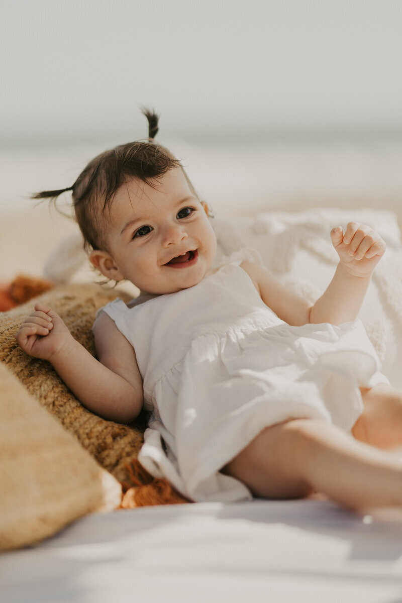 Petite fille en robe blanche avec deux couettes souriante, allongée sur des coussins à la plage capturée par Laura Termeau photographie en Vendée.