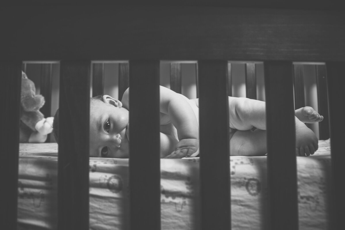 a black and white image of a newborn looking through the bars of its crib at the camera
