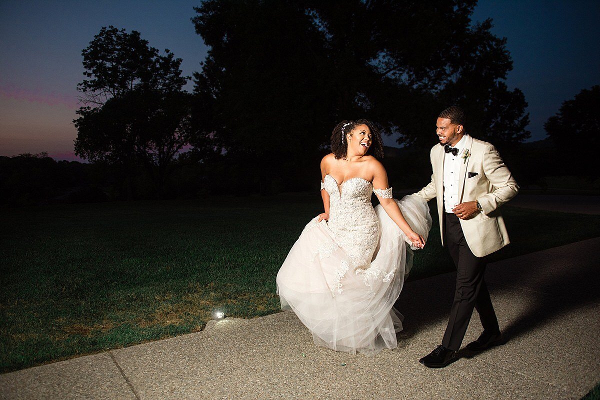 The groom, dressed in a black and white tuxedo, helps the bride with her long mermaid style lace wedding dress as they walk through the garden at Ravenswood Mansion after sunset