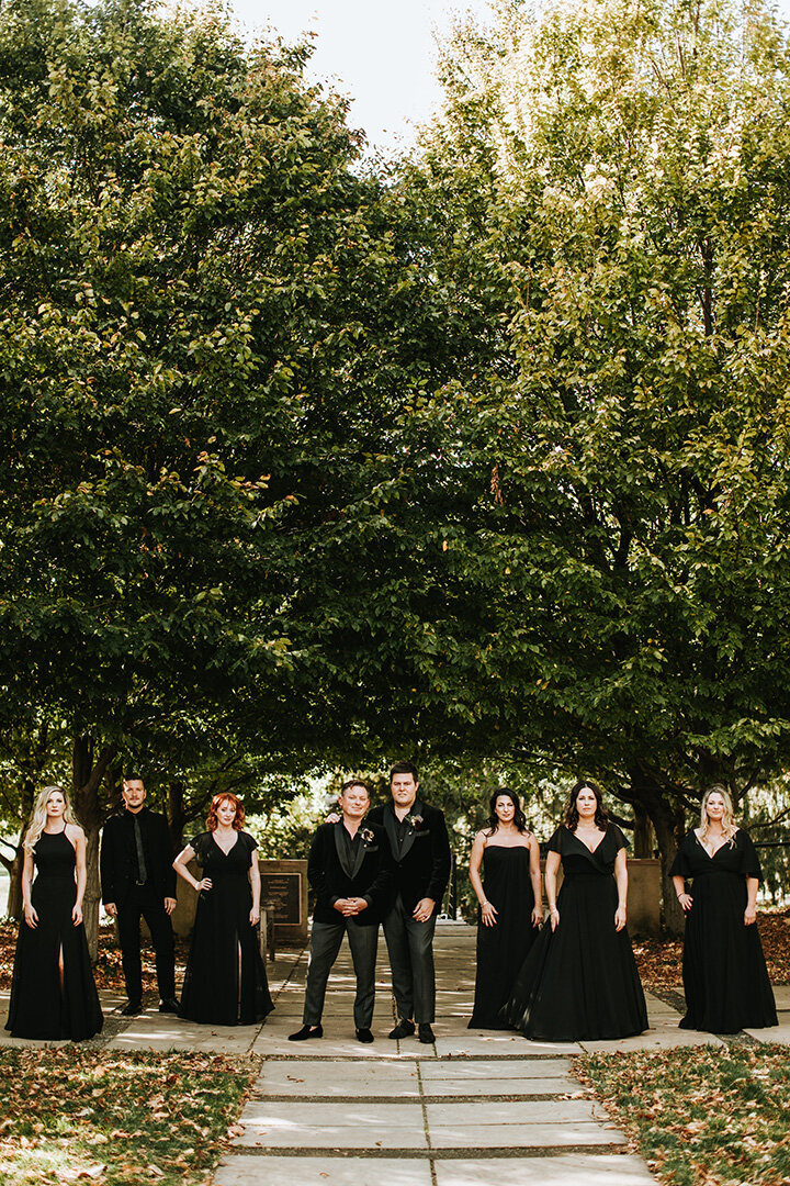 Wedding party wearing all black gowns and tuxedos pose outdoors, surrounded by large trees