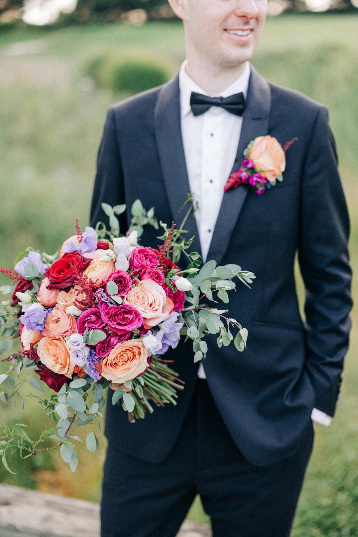 Groom holding a vibrant bridal bouquet, wearing a black tuxedo with a bow tie.