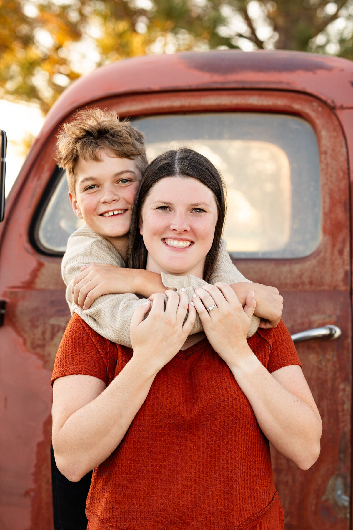 Preteen boy stands behind his mom and hugs her around the neck as they smile at the camera.