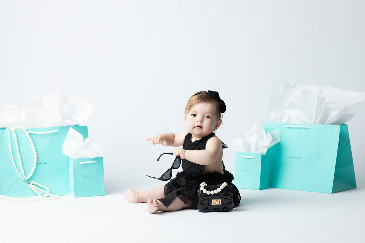 DFW milestone photographer highlights a baby girl dressed in a black lace outfit with a toy purse, pearls, and Tiffany gift bags, holding her sunglasses with a calm, poised expression.