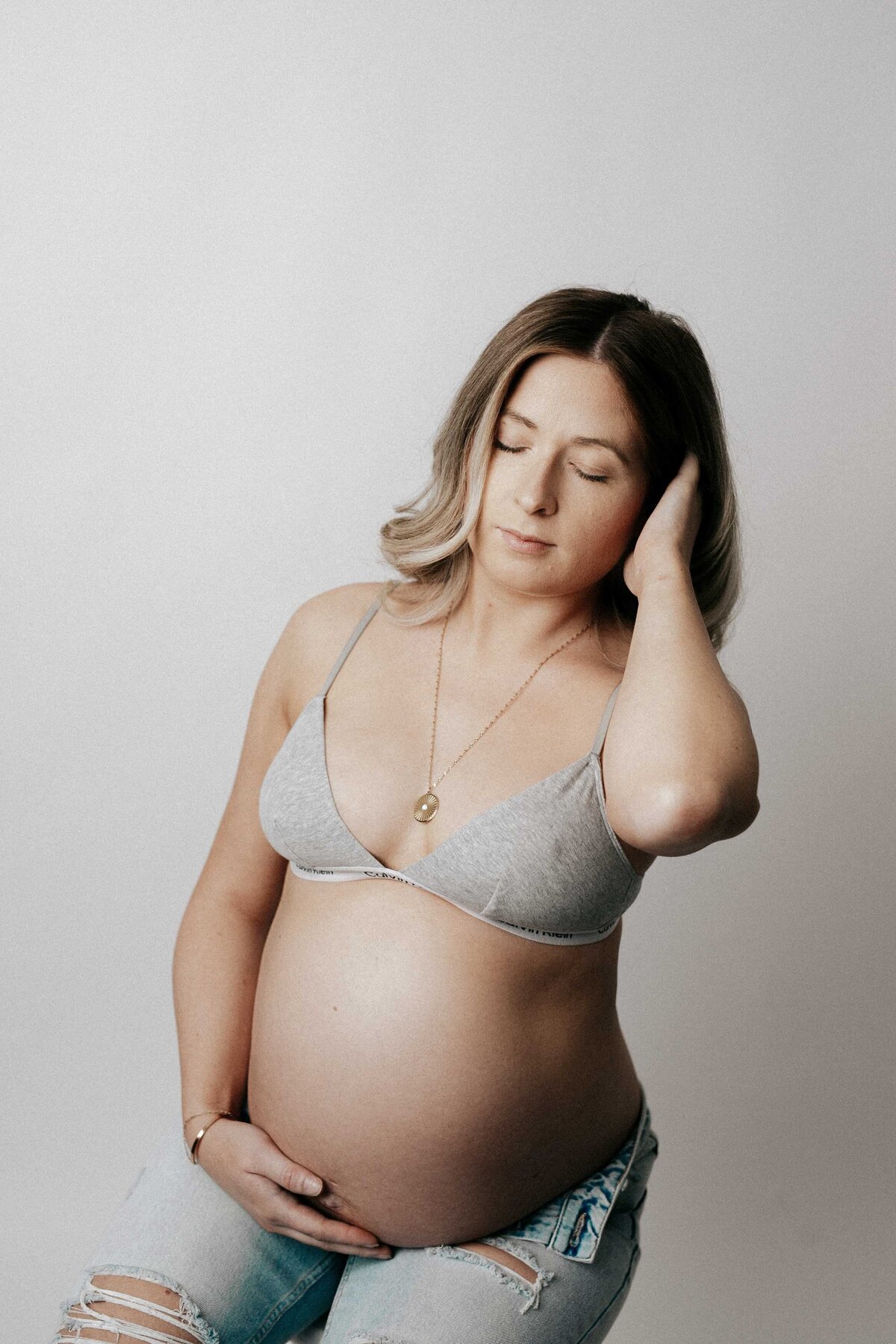 Studio maternity session. Mom is sitting on a stool with wearing distressed jeans and a grey bralette with her bump exposed. She is holding her bump and brushing the hair out of her face.
