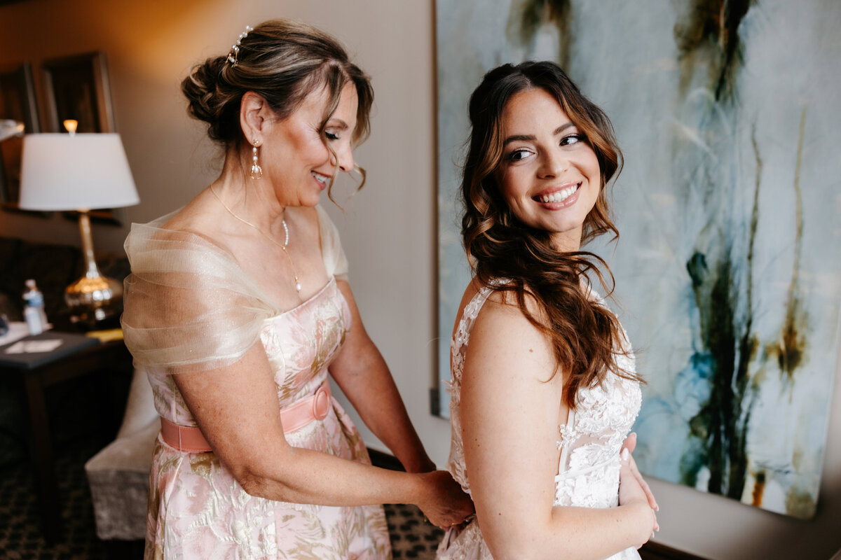 A bride with curly brown hair smiles back at her mom as she zips up her wedding dress,