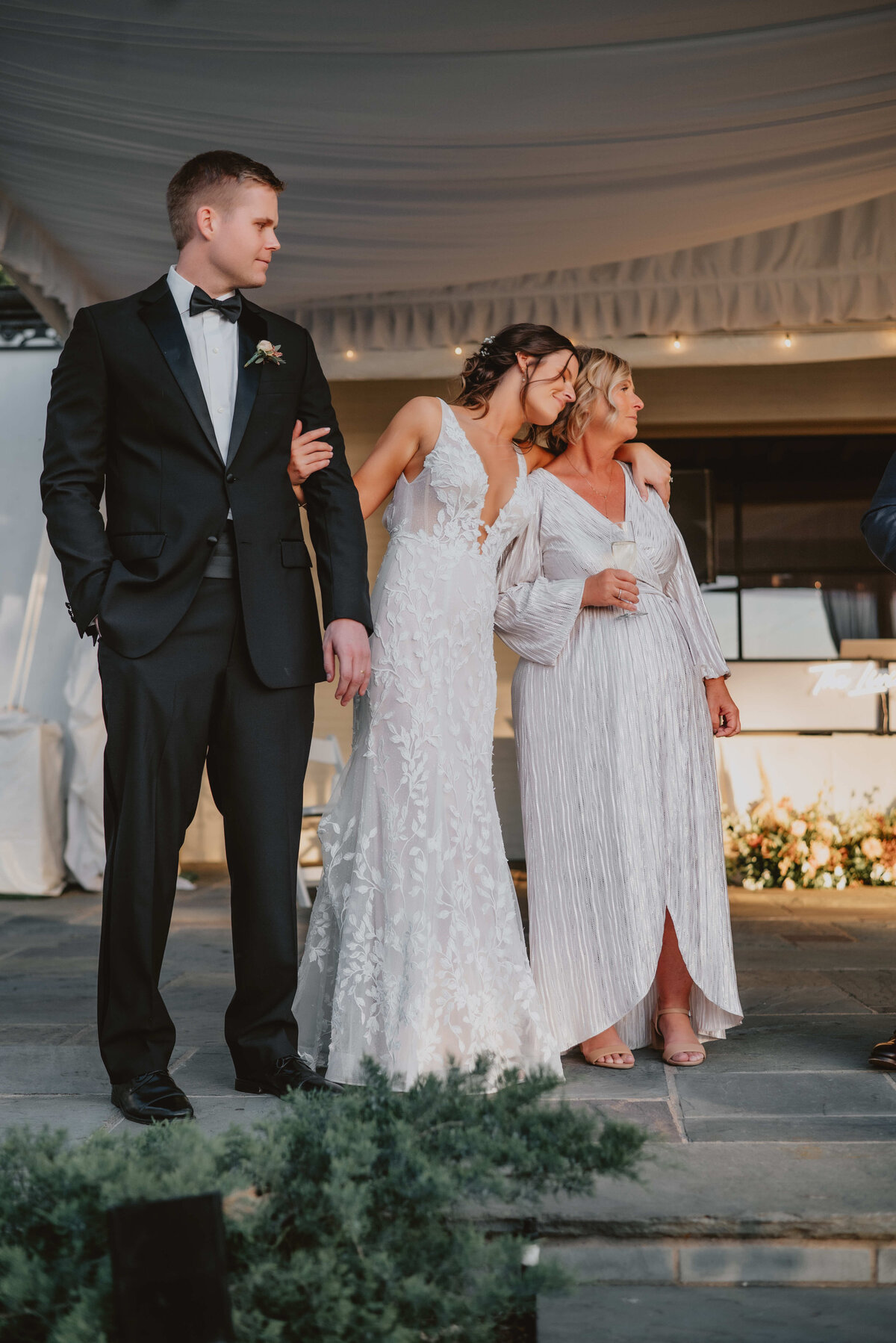 Bride in a lace gown embraces her mother in a silver dress while standing beside the groom in a black tuxedo during the wedding celebration.