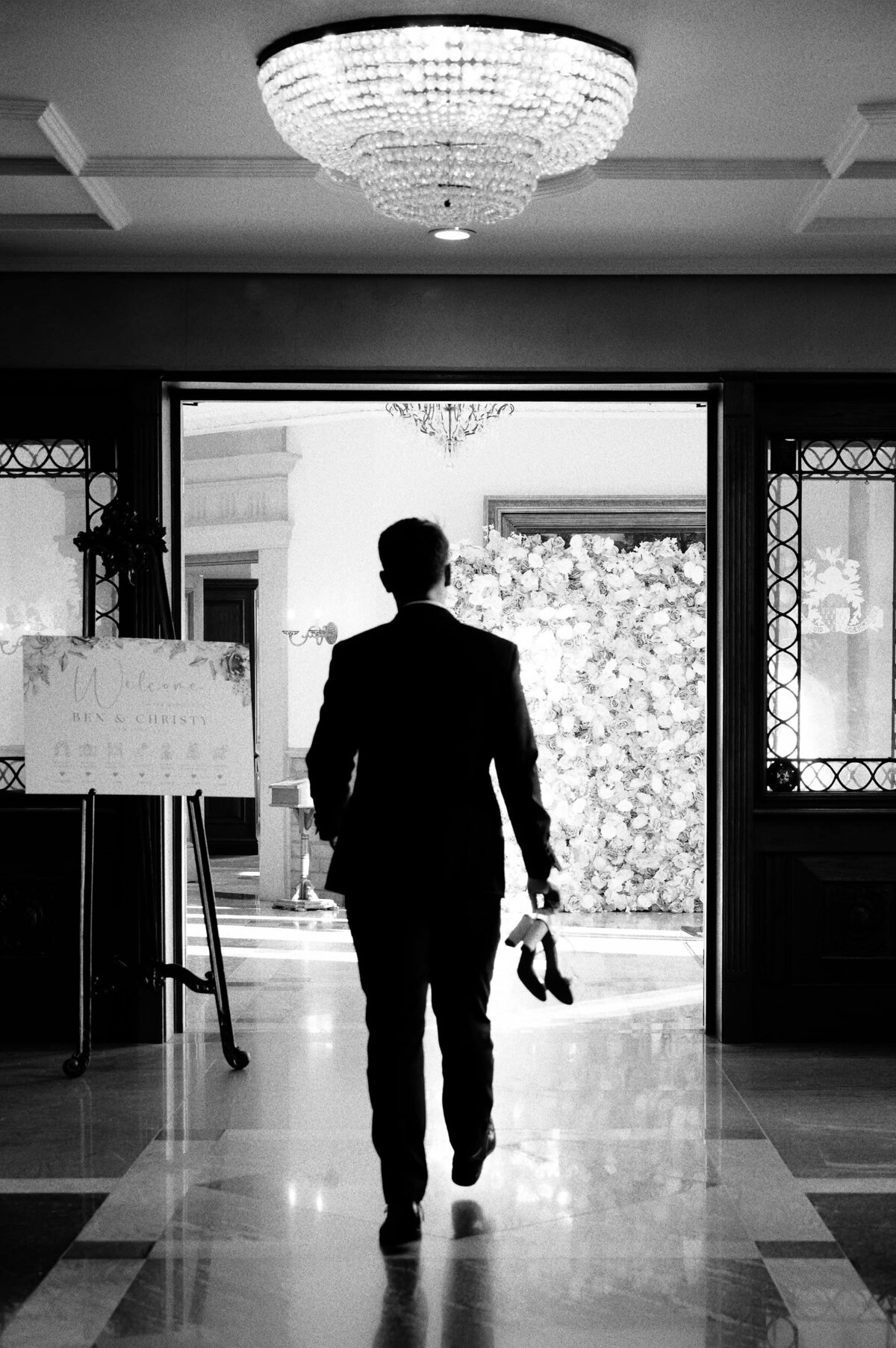 Black and white image of groom carrying his brides wedding shoes