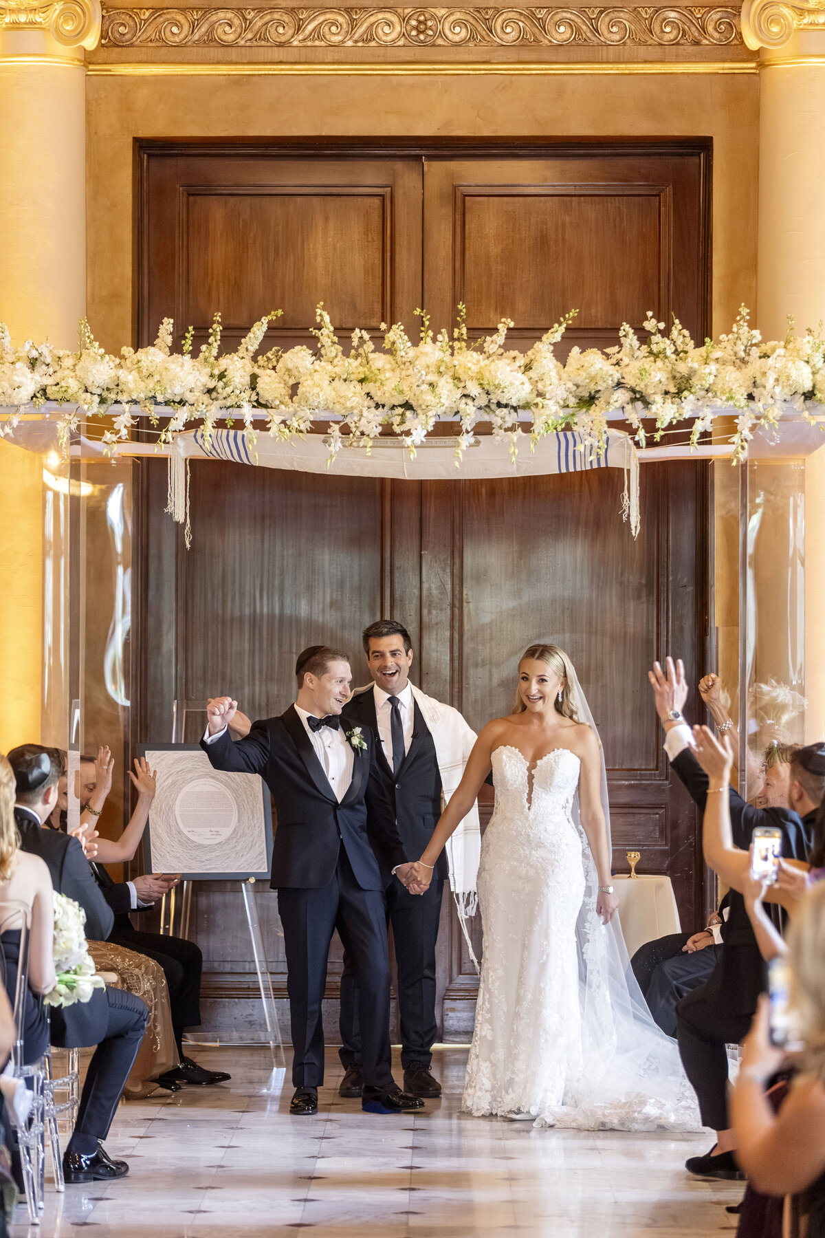 A joyful wedding ceremony with a couple holding hands under a floral arch. The groom is in a suit, and the bride is in a strapless white gown. A man stands beside them, smiling. Guests are seated, some raising their hands in celebration.