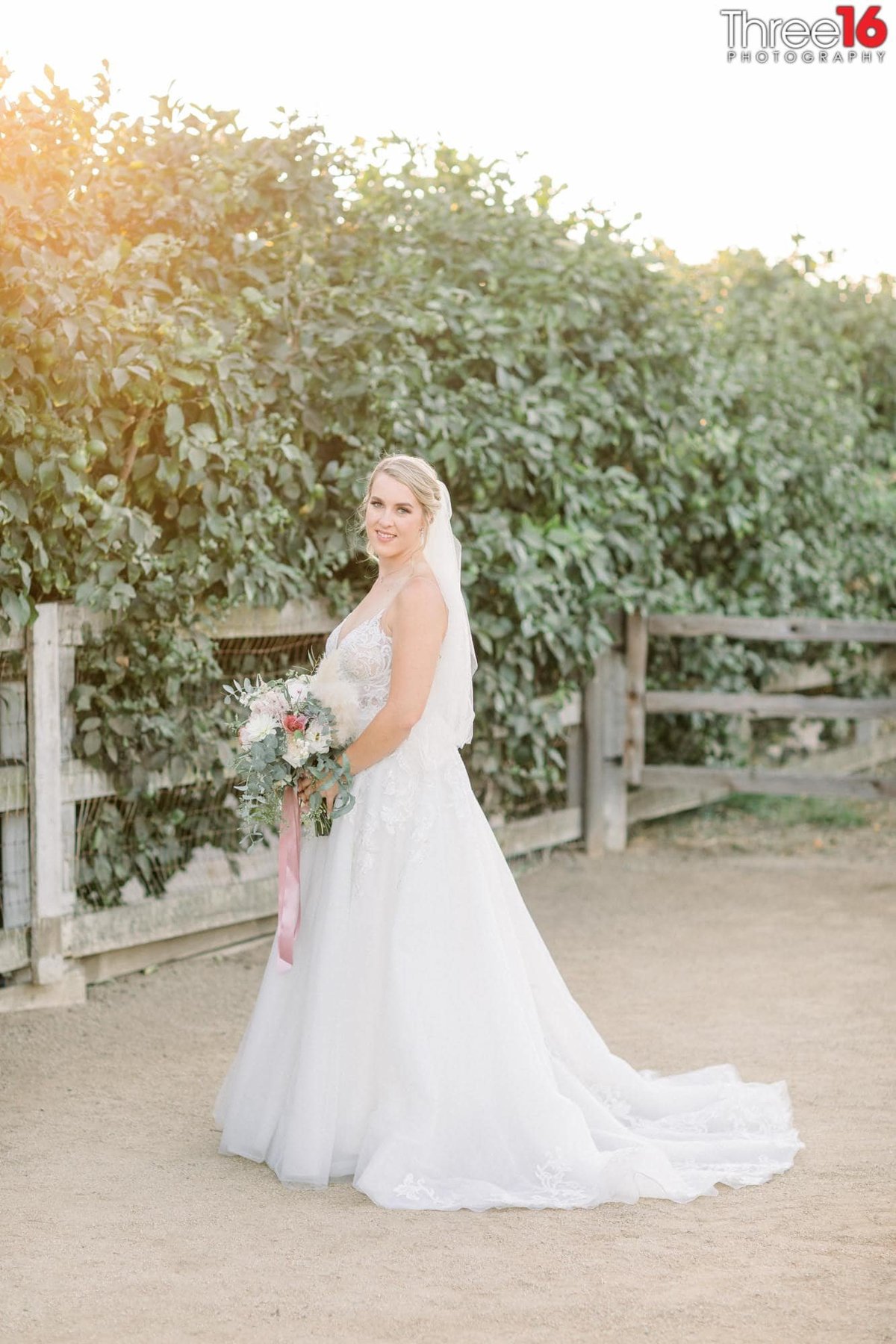 Bride poses with with her bouquet and dress train fanned out in front of a ranch-style fence