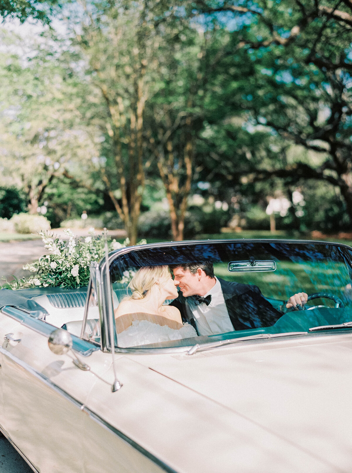 Newport Wedding Photographer | A couple in wedding attire shares a joyful moment in a vintage convertible. Sunlight filters through trees.