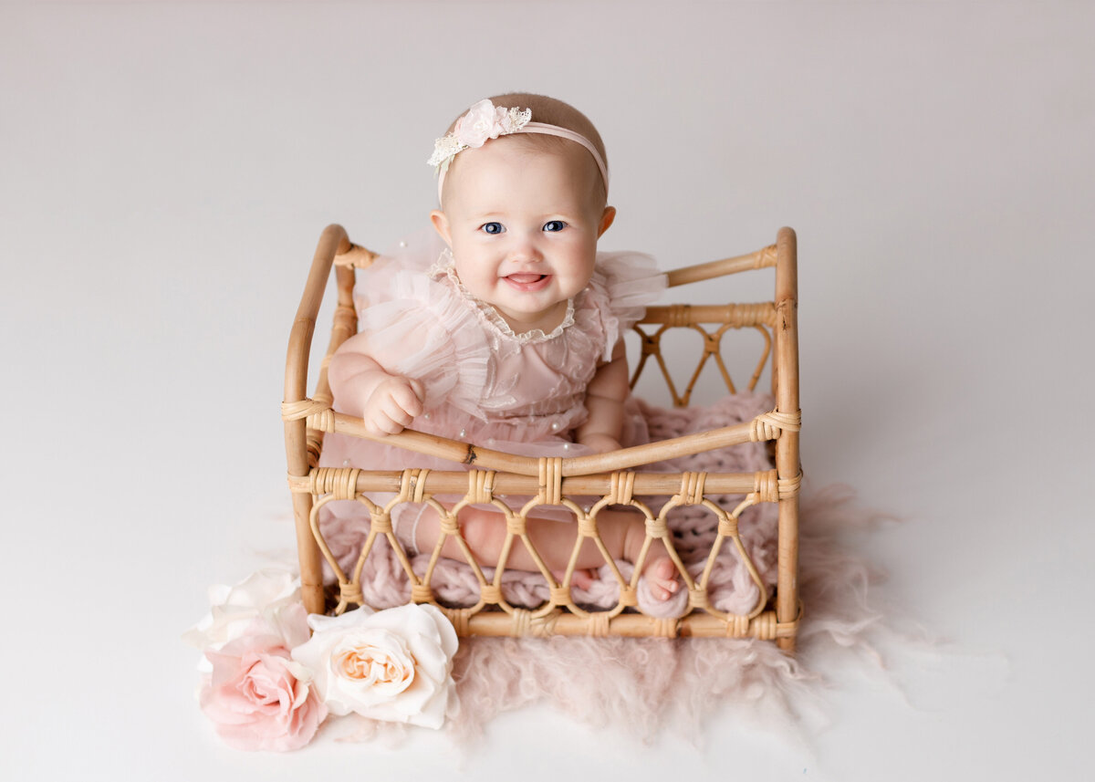 Baby girl sitting in a miniature vintage-inspired crib. Baby is wearing a pink dress and matching headband. Baby is looking up smiling at the camera.