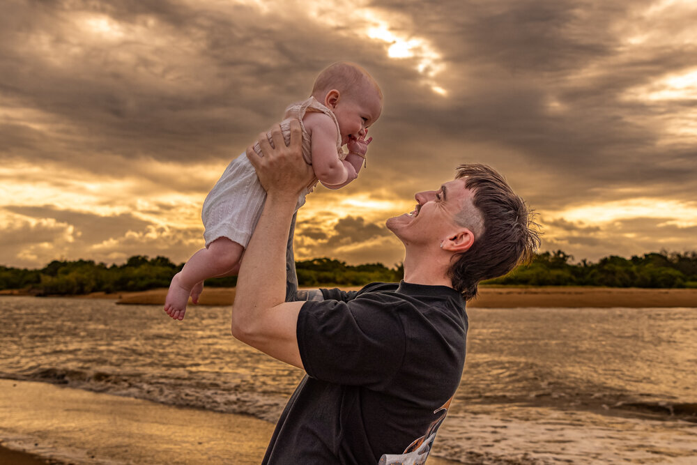 new dad holding baby girl into the air at the beach at sunset - Townsville Child Milestone Photography by Jamie Simmons