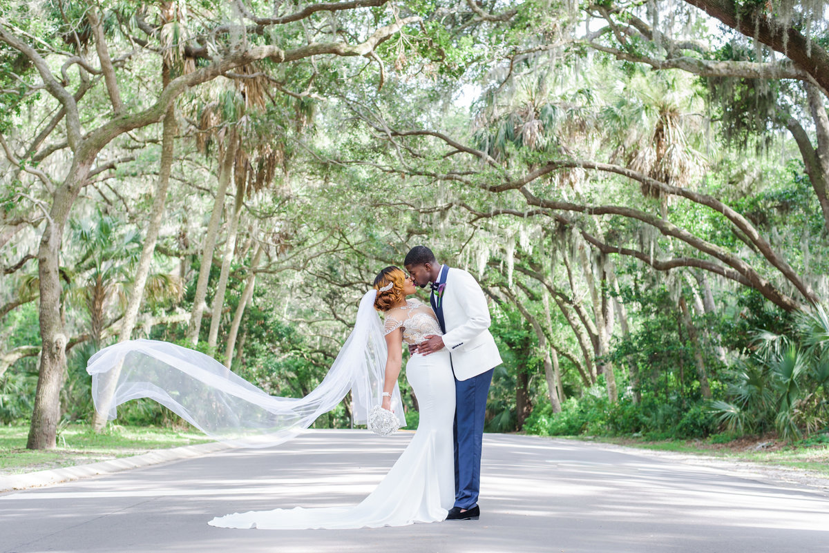 Bride and Groom kiss under an arch of oak trees as her veil blows in the wind in Dunedin, Florida