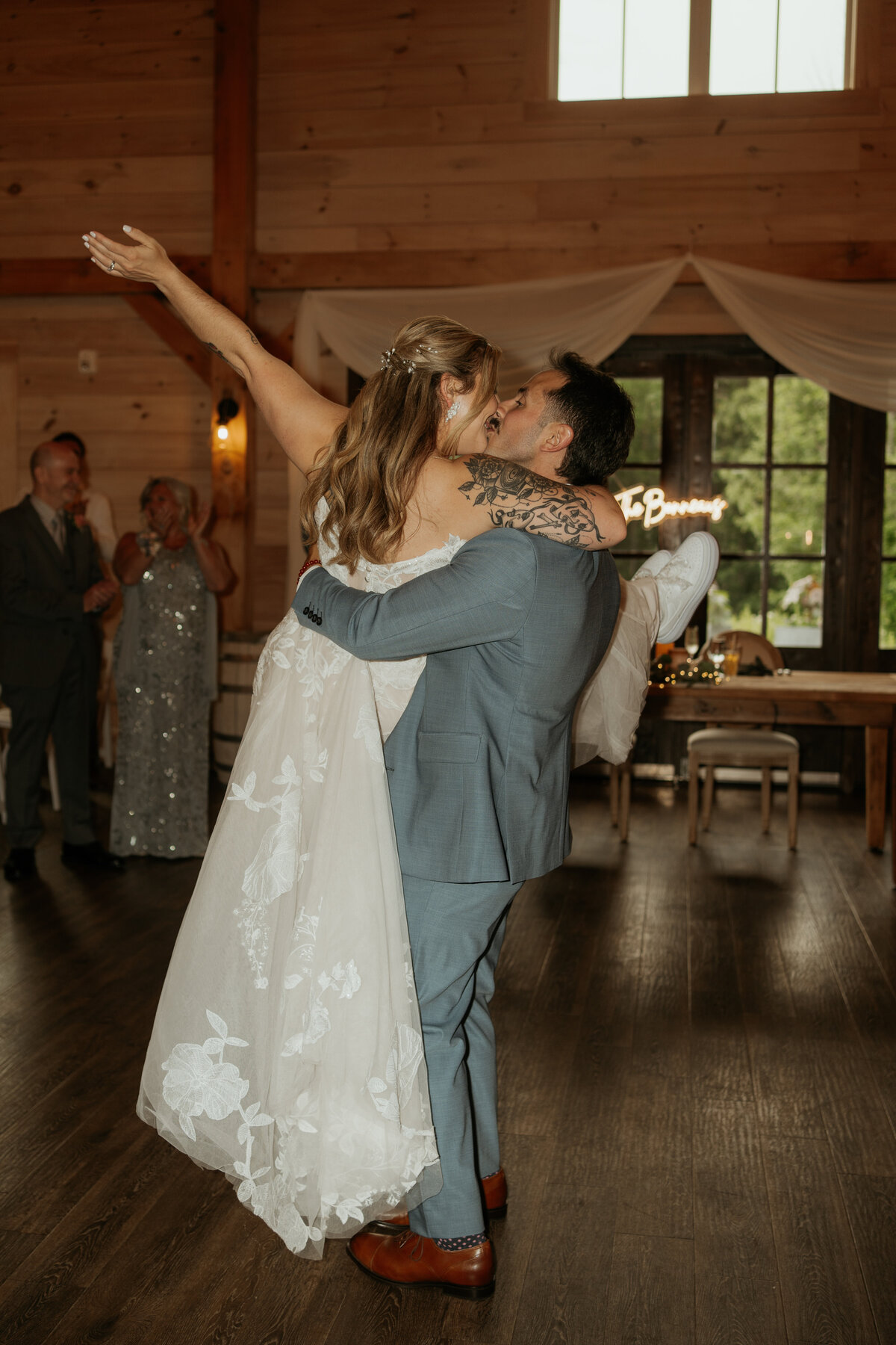 Bride and groom share an enthusiastic embrace on the dance floor during their wedding reception. The bride, in a lace gown with visible tattoos, lifts her arm in celebration while the groom, in a light grey suit, holds her tightly. Guests in the background applaud and cheer, highlighting the joyous and lively atmosphere of the occasion.