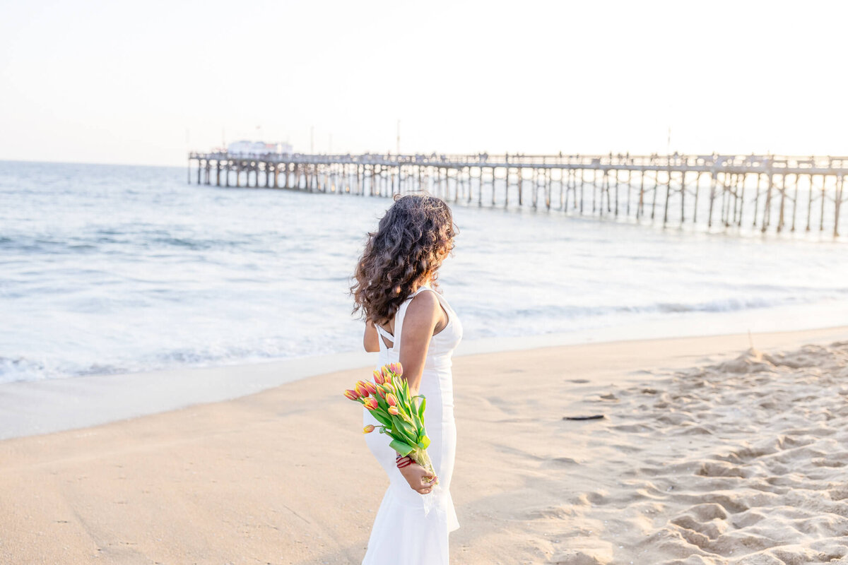 A woman in a white dress stands on a sandy beach, holding a bouquet of flowers. The ocean waves gently lap the shore, and a long pier extends into the water under a bright sky.