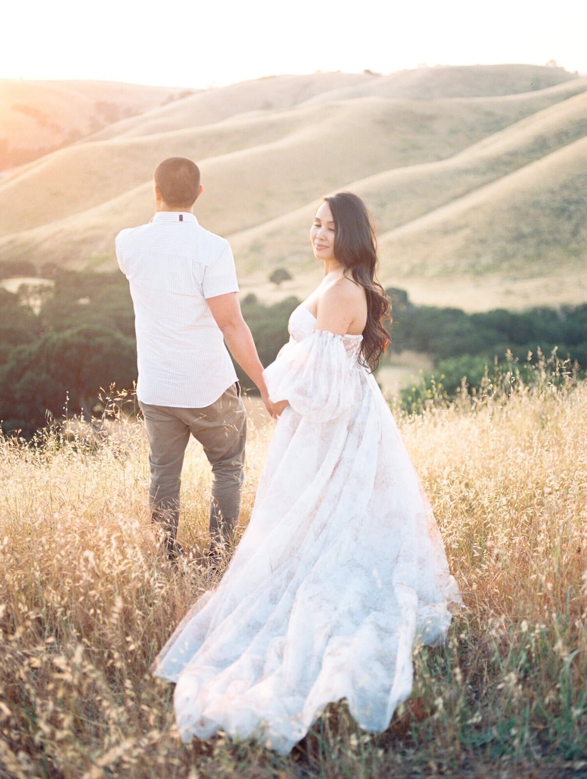 Maternity photoshoot in San Francisco with a pregnant woman and expecting dad standing in a field against hills.