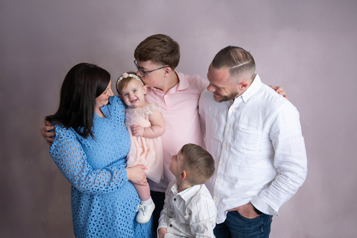 Smiling child surrounded by family, with big brother kissing her gently on the back of her head as mom holds her.