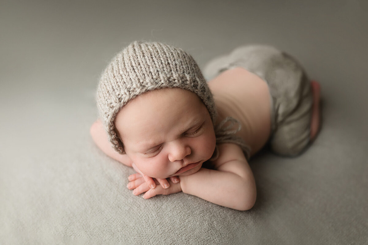 Newborn boy posed on gray back drop wearing gray pants and bonnet.
