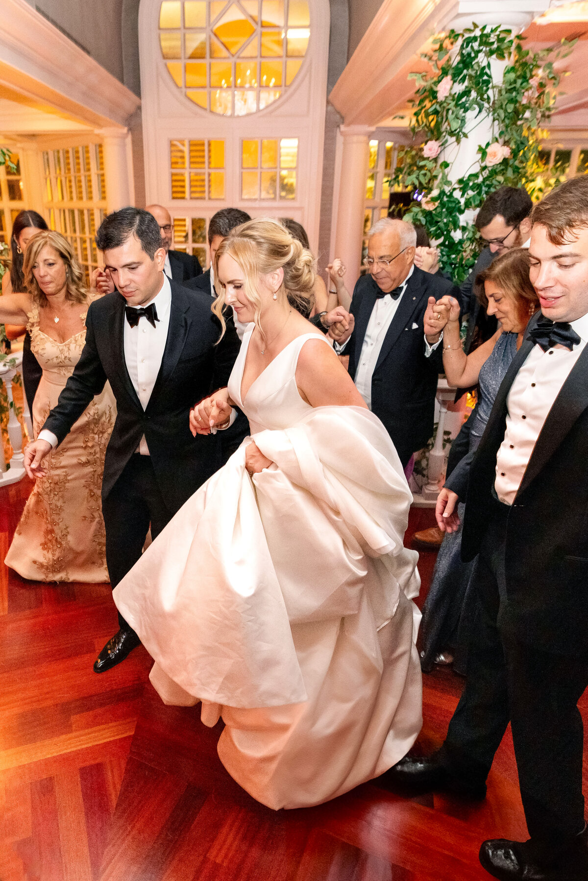 A bride in a white gown dances with a group of people in formal attire at an indoor event with wooden floors and elegant decor. They are holding hands and moving joyfully in a circle.