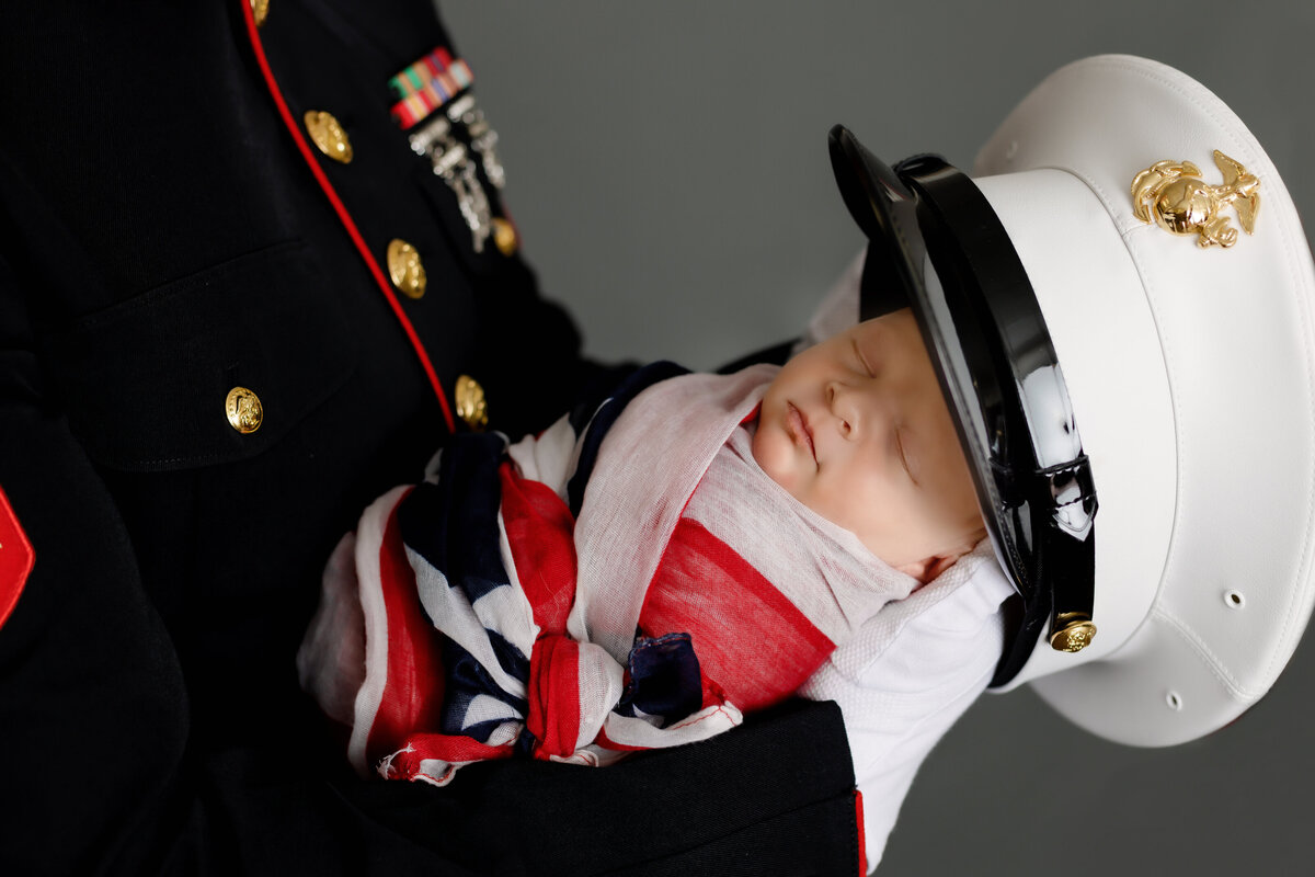 Newborn Photographer, a marine holds a baby wearing a marine hat`