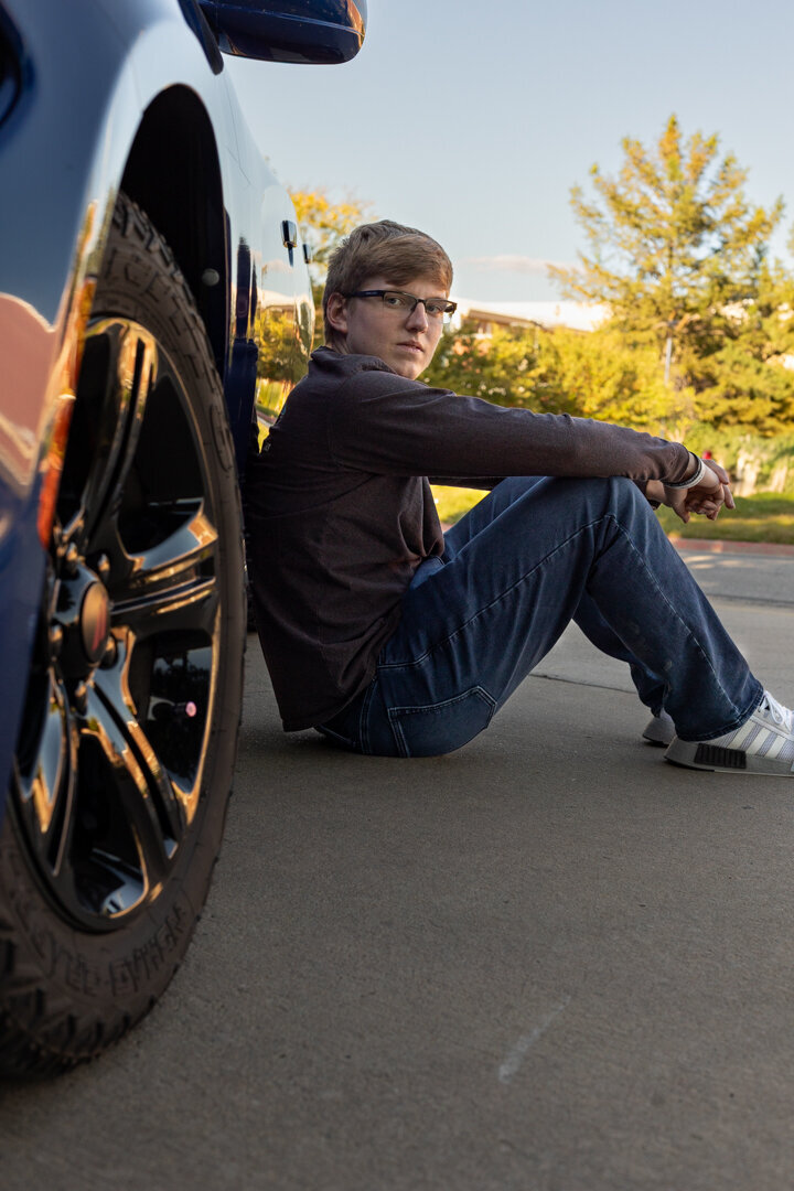 A senior guy sitting on the ground leaning against his car in Lawrence, KS