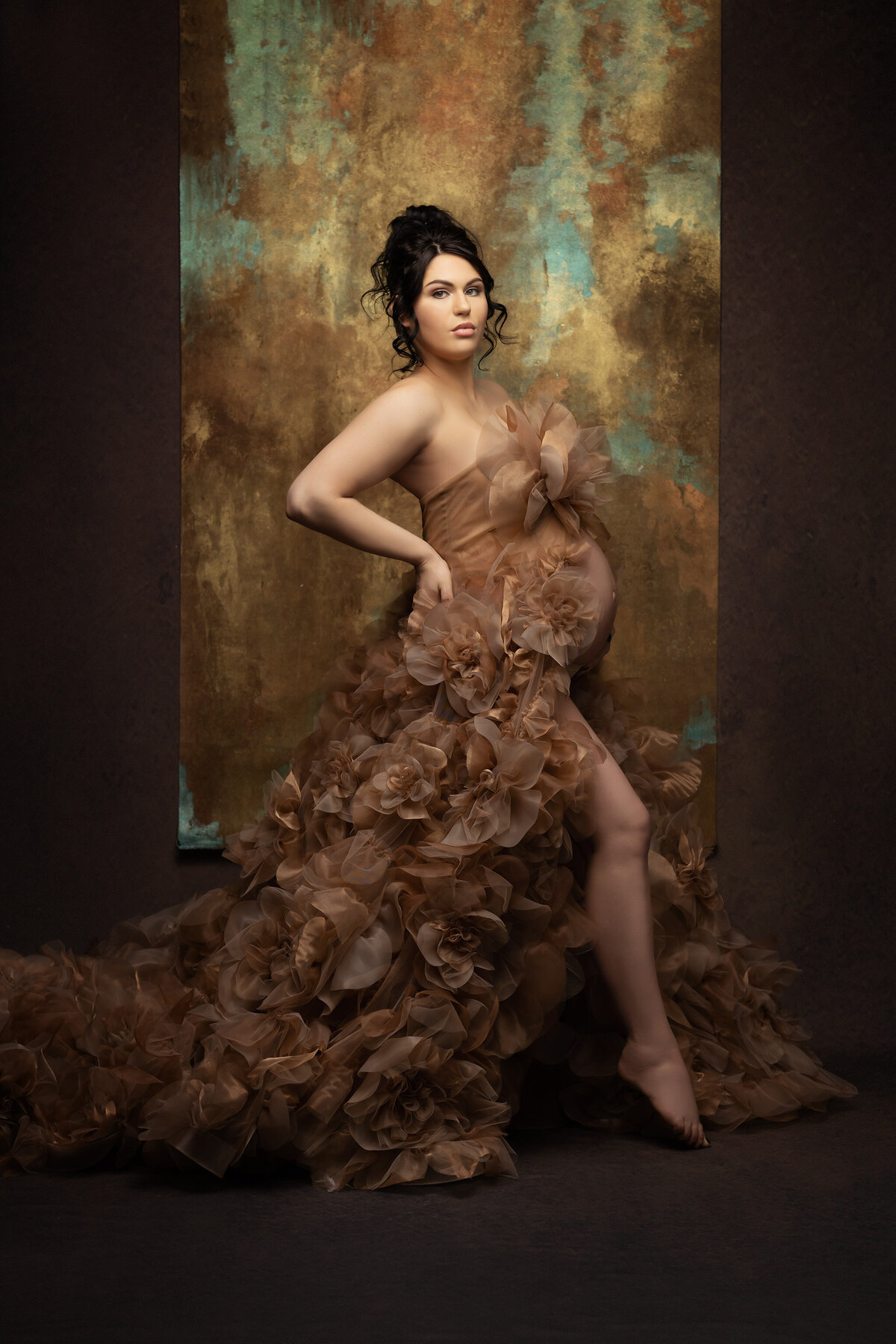 Pregnant woman in a flowing, floral gown against a textured gold and teal backdrop.