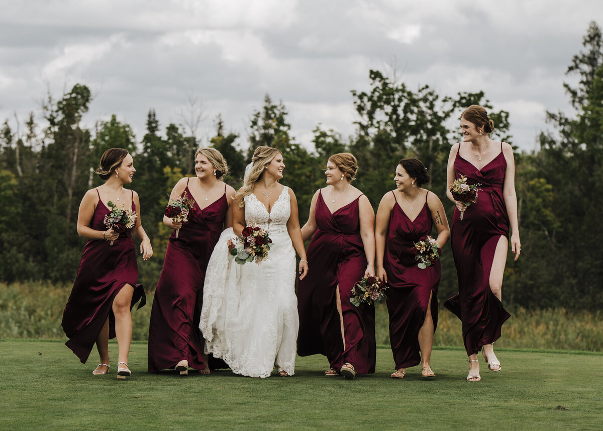 A joyful bride in a white gown shares a moment with her bridesmaids, who are adorned in elegant maroon dresses, as they walk across a verdant field with a hint of overcast skies above taken by jen Jarmuzek photography a Minneapolis wedding photographer