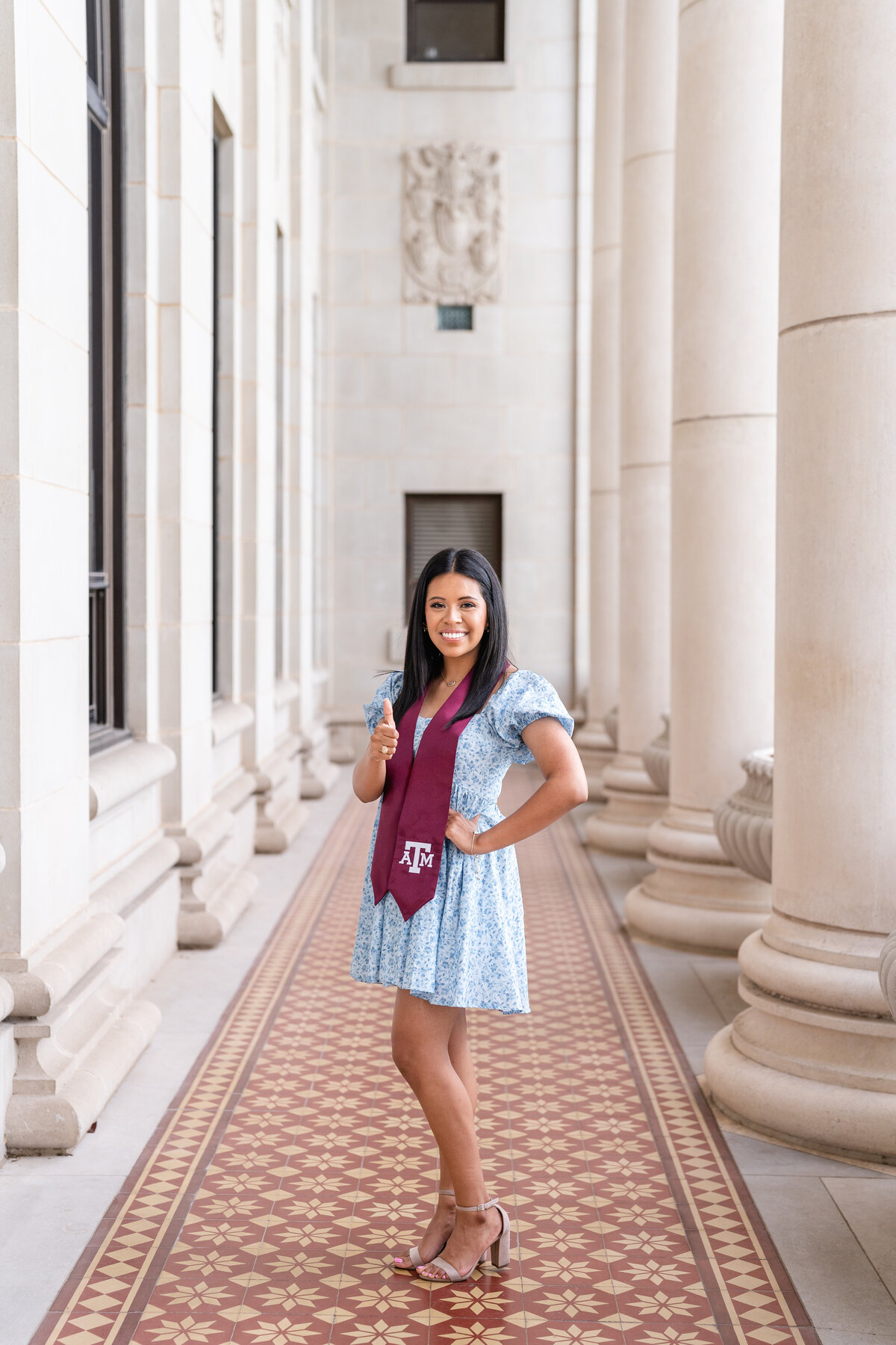 Texas A&M senior girl standing with hand on hip and thumbs up wearing blue dress and maroon stole in columns of Administration Building