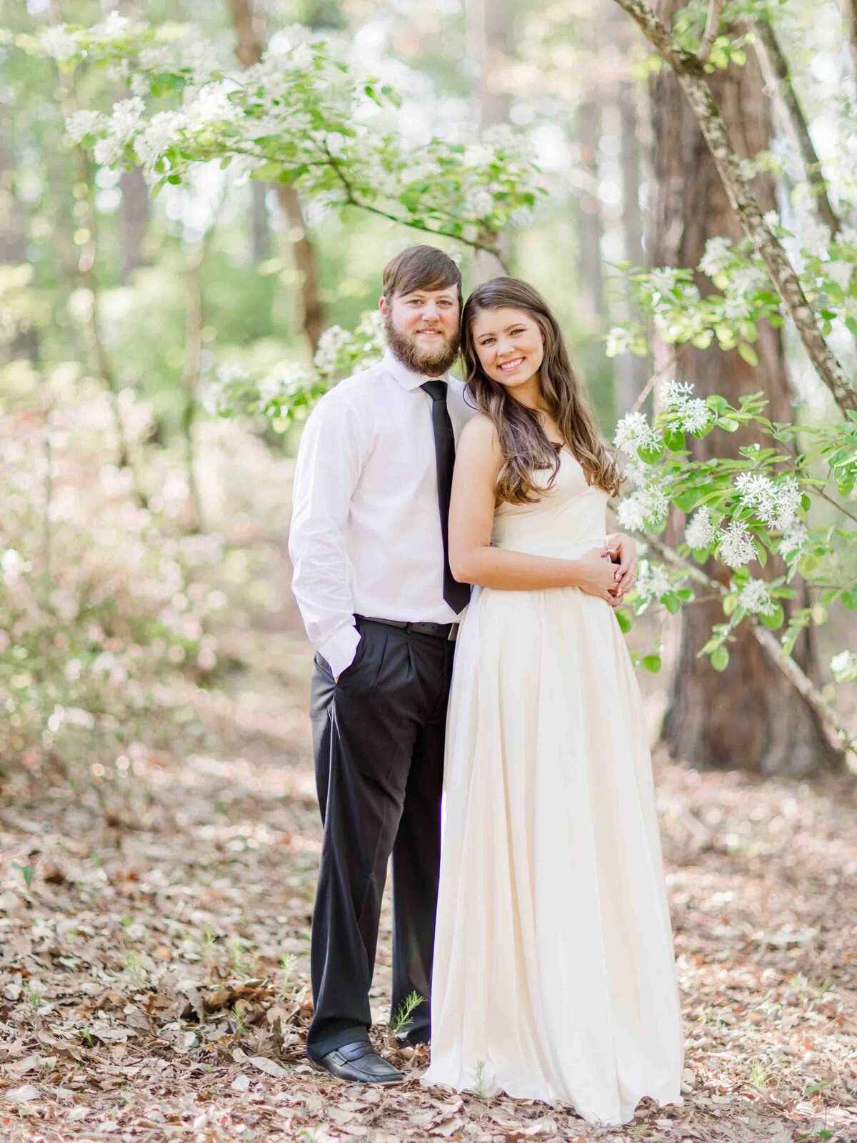Engagement portrait of a couple in the forest with flowers while she wears a white dress and he wears a suit.