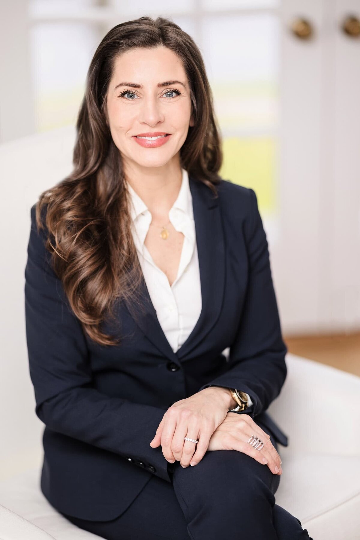 Professional headshot of a woman with long brown hair in a navy suit, seated indoors on a white chair in Long Island.