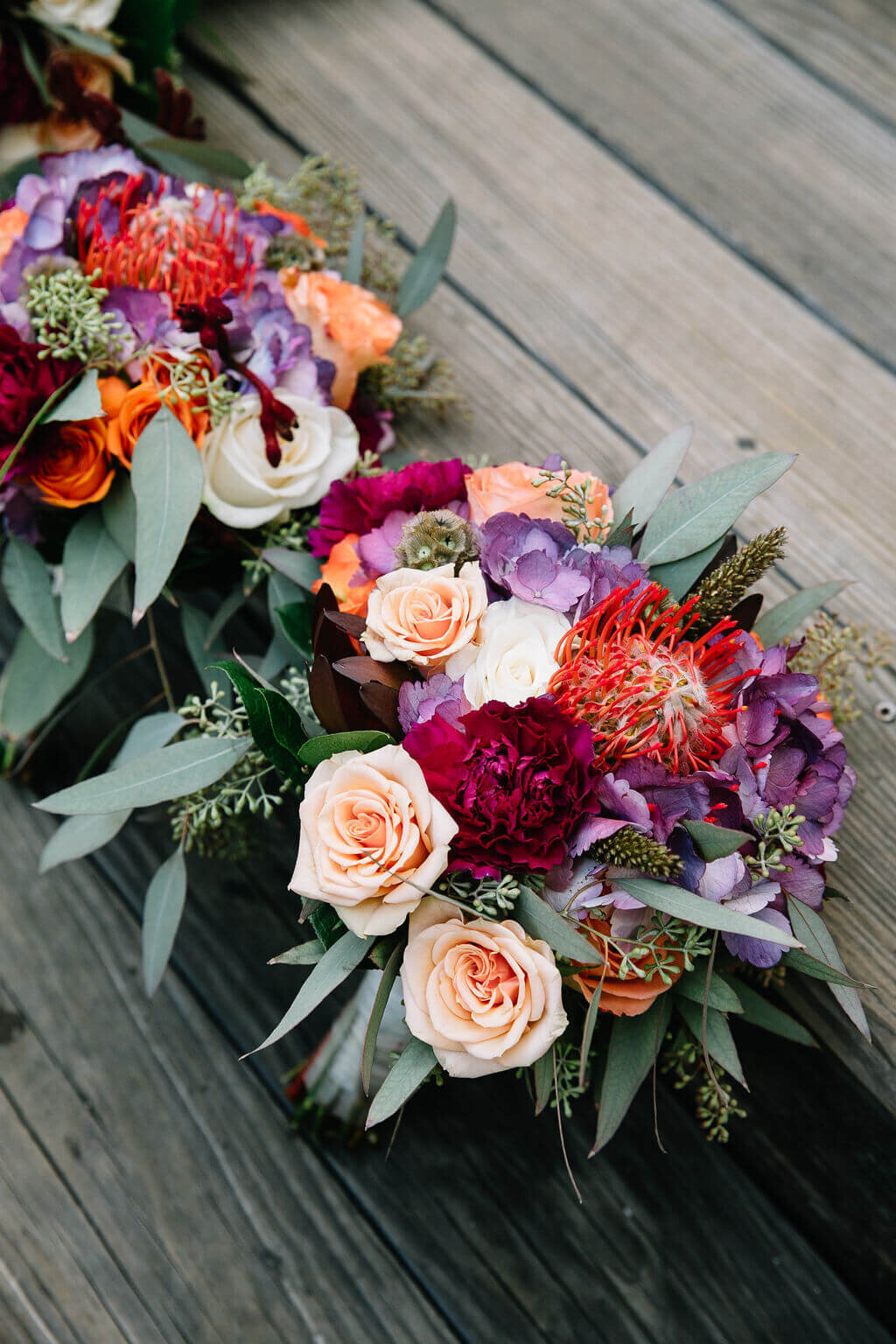Several vibrant floral bouquets featuring roses, eucalyptus, and other colorful flowers are lined up on a wooden deck.