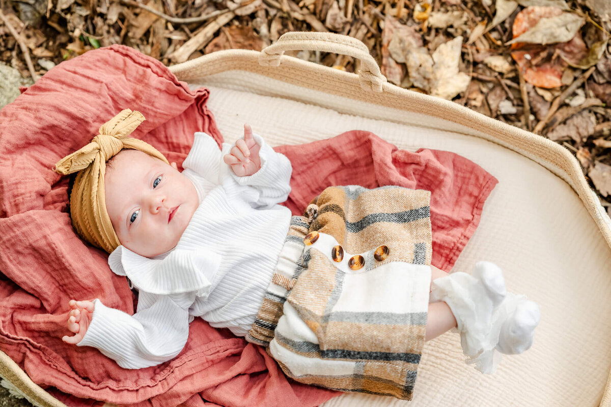 A baby girl wearing a plaid skirt and mustard color bow lays on top of a red blanket in a moses basket. Photo by Justine Renee Photography near the Outer Banks.
