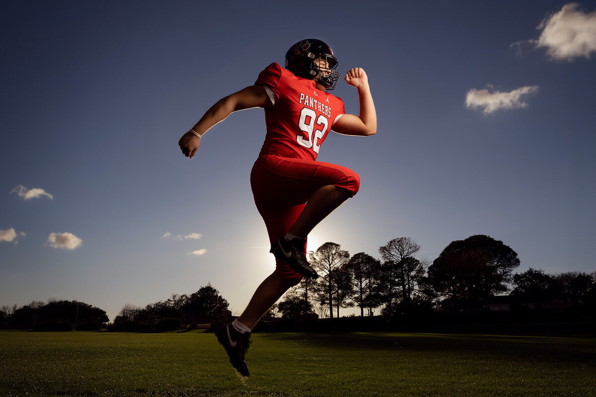 Dynamic senior portrait of a football player captured mid-action, showcasing powerful motion during a jump. The vibrant red football gear contrasts with the dramatic lighting, highlighting the energy and determination of the athlete. This Dallas senior photography session embodies strength and movement, perfect for those looking for a unique, sports-inspired senior photo. The intense sky and glowing sunlight add to the visual impact, creating a truly memorable portrait.