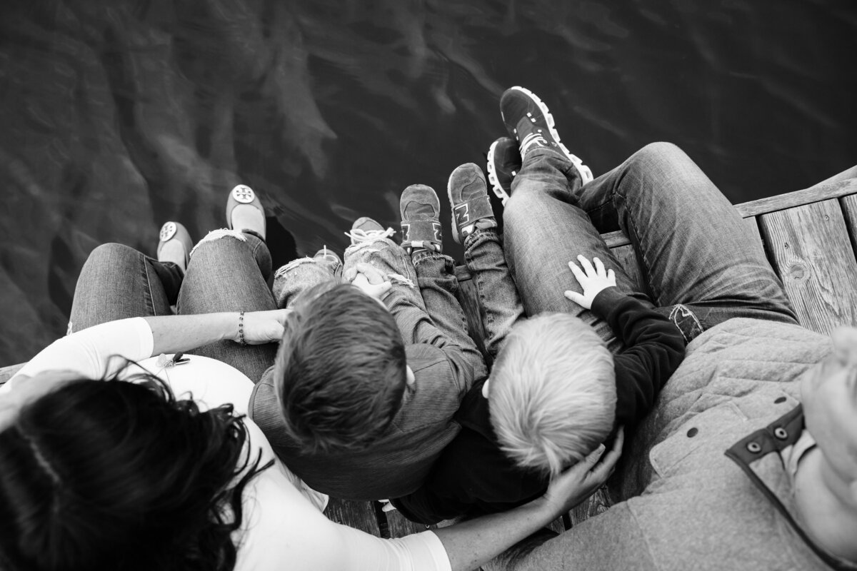 A top-down view of a family sitting on a dock, with their feet hanging over the water. The parents sit with their two children nestled between them, capturing a serene family moment.