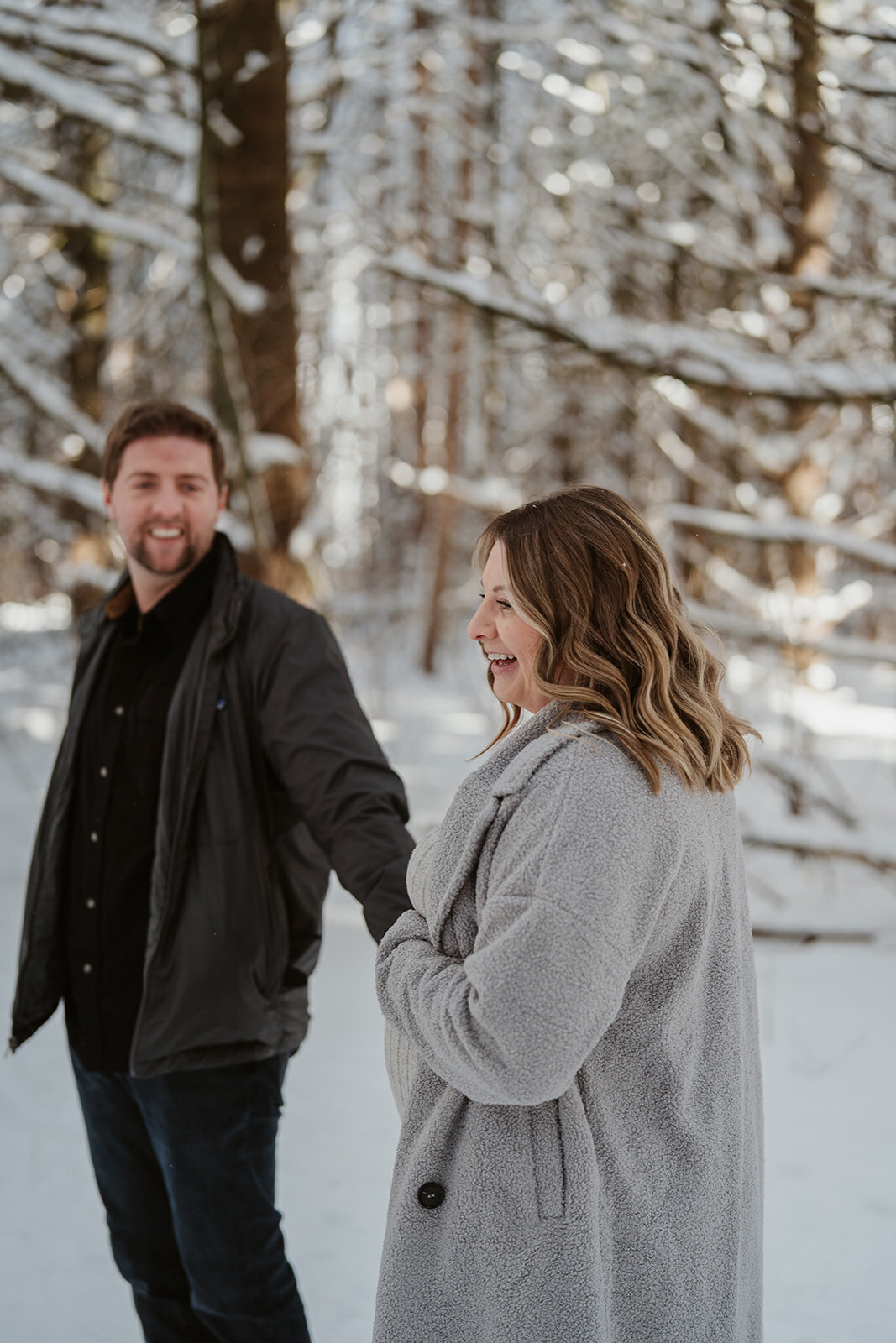 Expecting couple smiling and walking together through the snowy woods at Al Sabo Preserve during their maternity photoshoot.