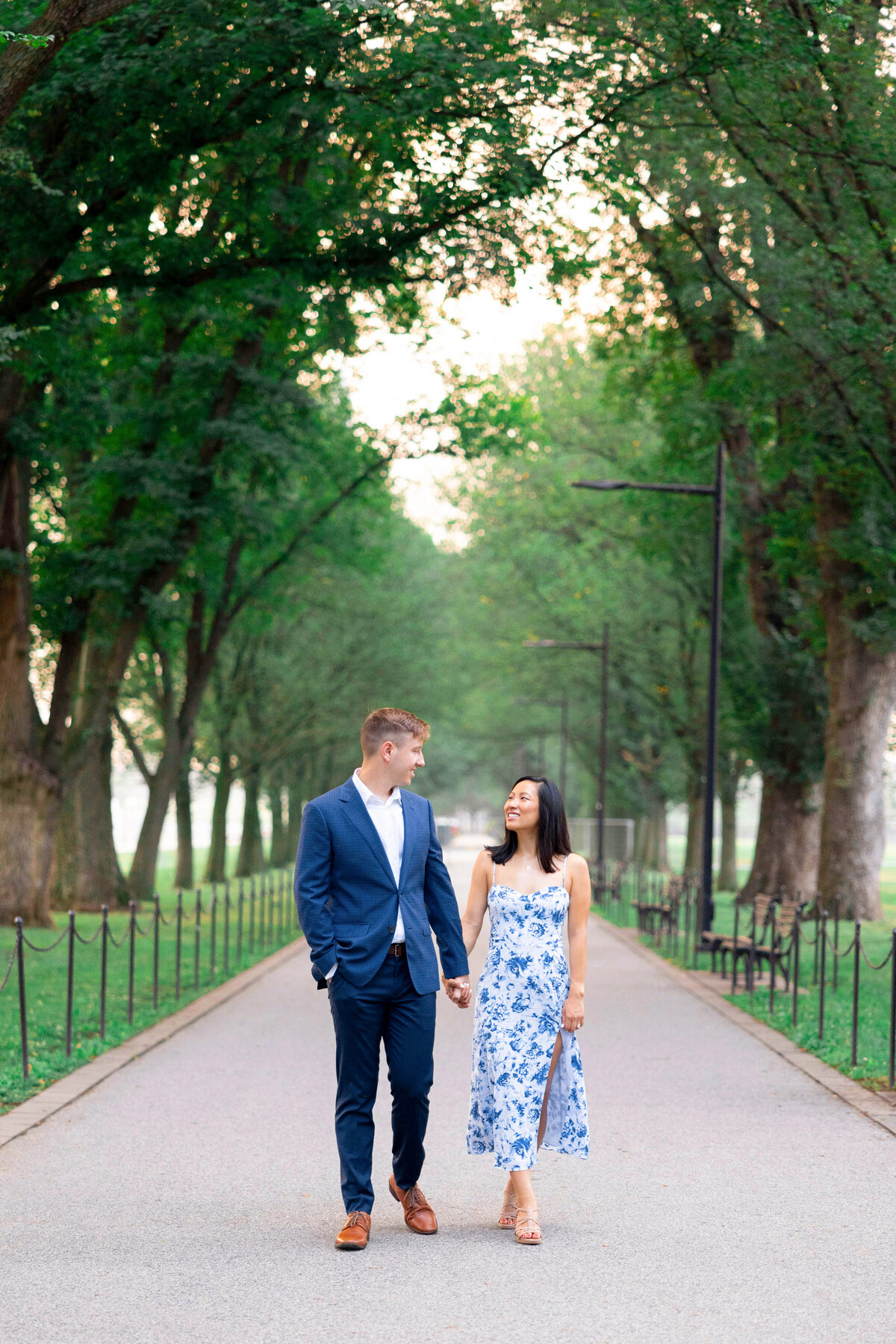 A couple holding hands walks down a tree-lined path. The man wears a blue suit, and the woman wears a white and blue floral dress. They smile at each other, surrounded by greenery and soft, natural light.