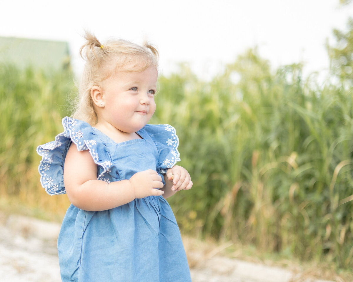 Toddler girl with blonde pigtails, blue and white dress, standing in rocked path at a wildlife sanctuary, looking away from the camera with a soft smile
