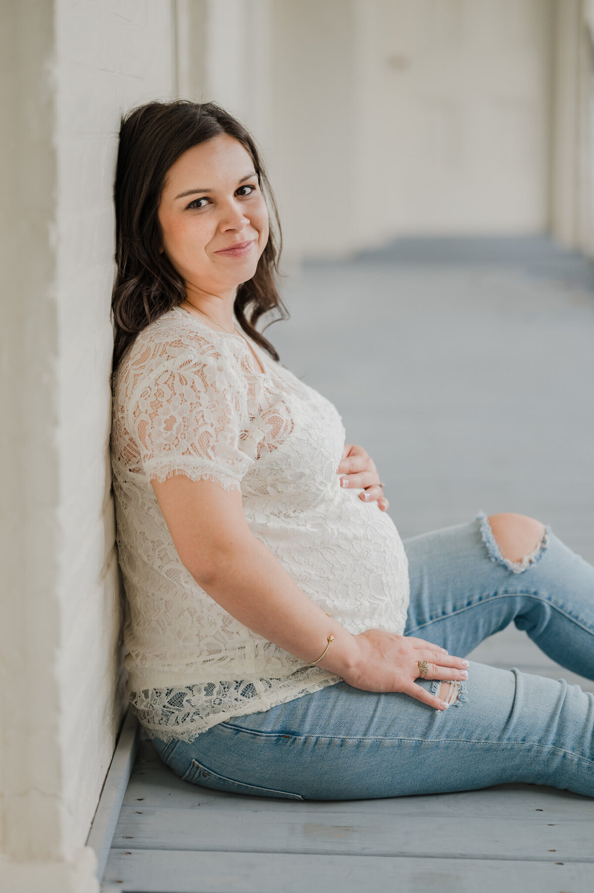 Pregnant brunette in jeans and a lace shirt sits and smiles at the camera during maternity pictures.