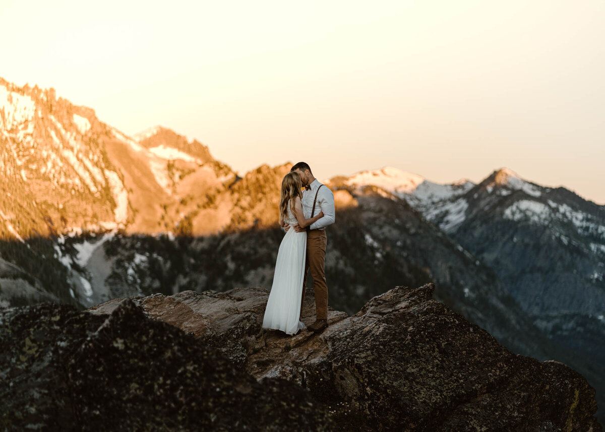 after learning how to elope in washington state, a couple kisses on top of a mountain with snowy peaks behind them