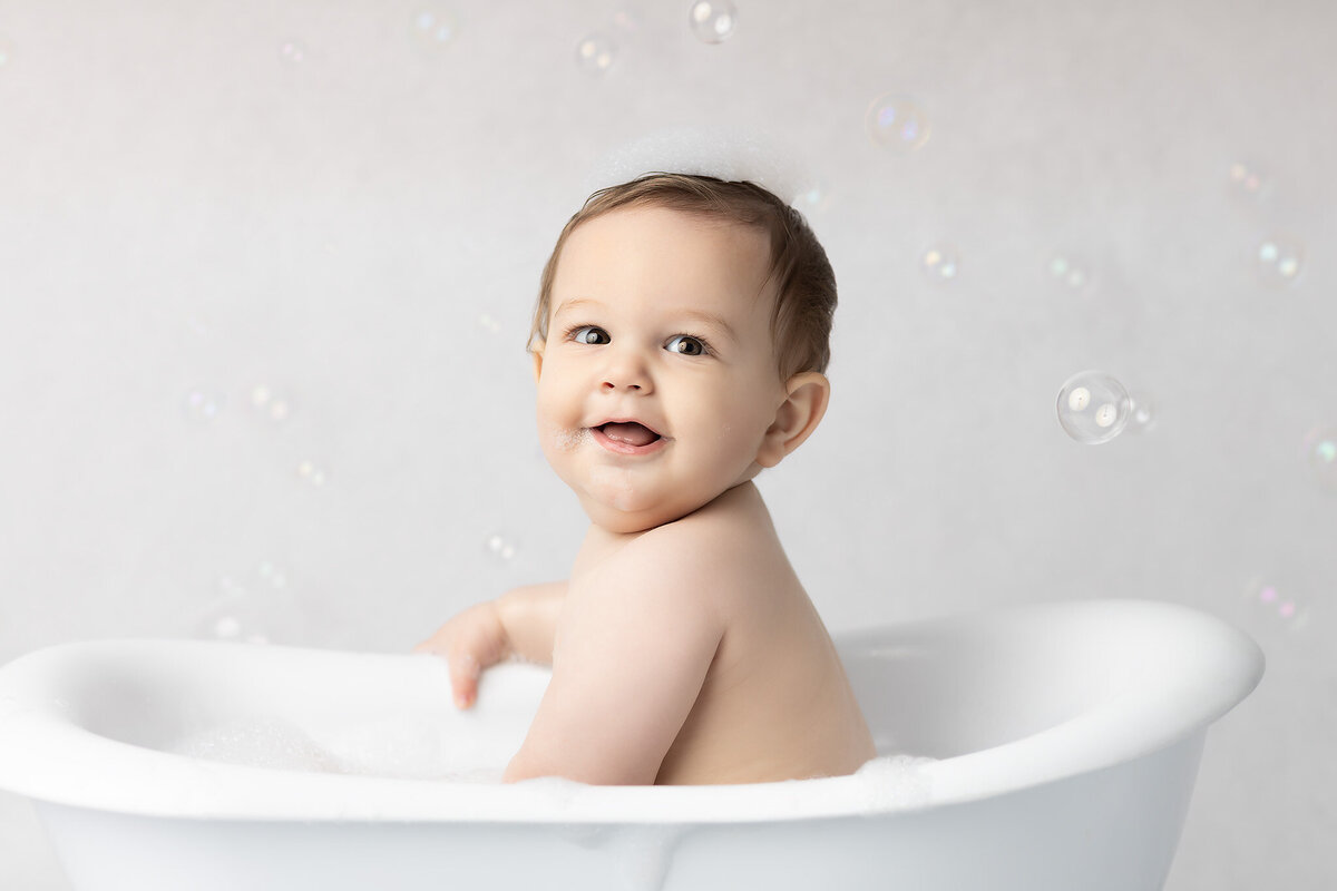 Dallas milestone photographer captures a happy baby with bubbles on their head, leaning over a white tub and smiling with joy during a bubbly bath session.