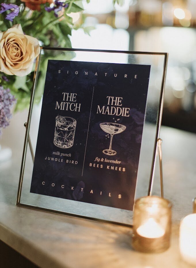Close-up of a gold-framed signature cocktail menu featuring "The Mitch" and "The Maddie," designed in a navy blue modern style with line art illustrations of each drink. Beside the frame, vibrant florals and a candle add a cozy touch. This elegant menu design, displayed at a wedding in North & Navy, Ottawa, perfectly balances personal style with the venue's old-world Italian charm, creating a memorable cocktail experience for guests. Ideal inspiration for couples seeking unique and stylish wedding stationery.