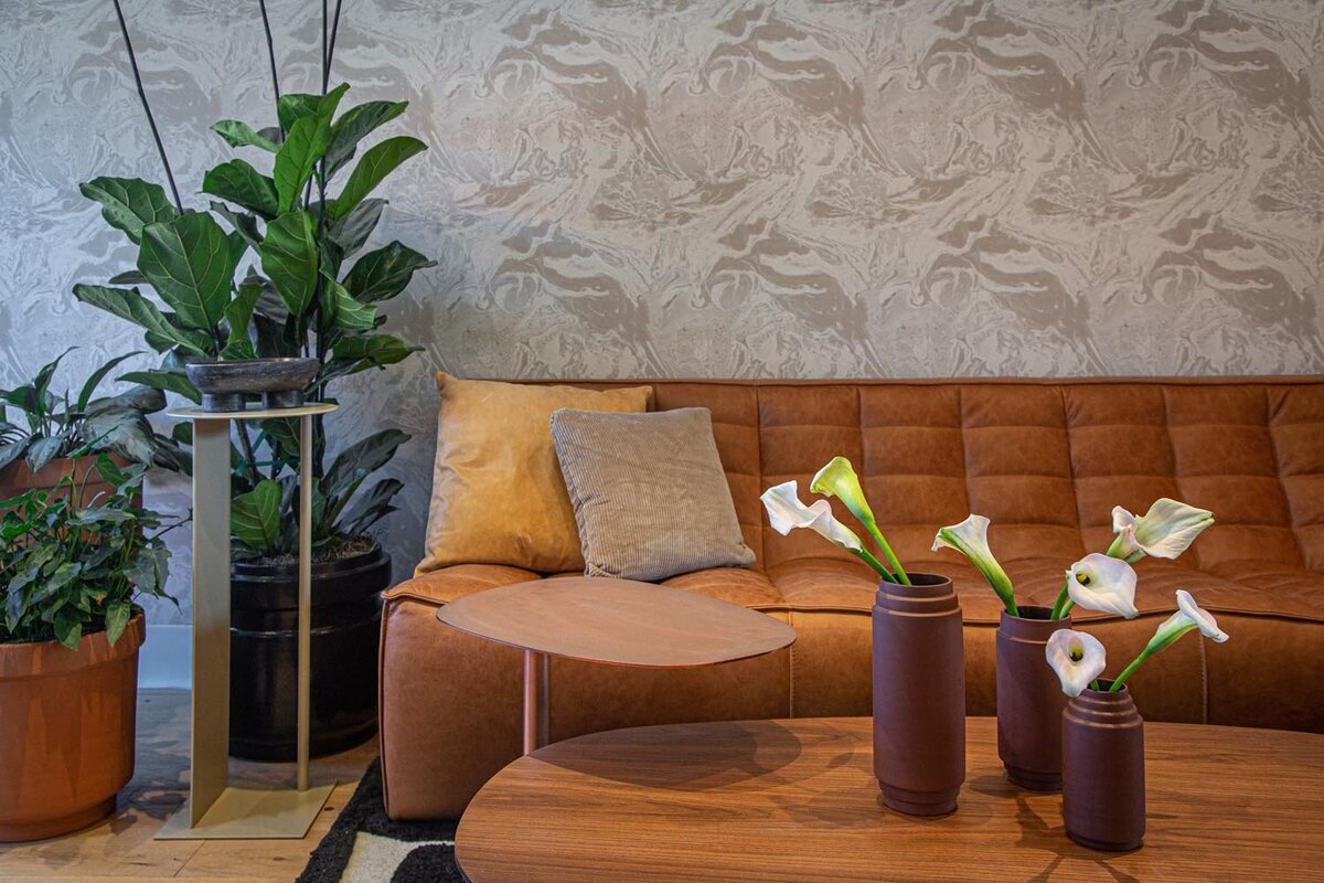 Seating area with brown leather sofa, patterned wallpaper, and ceramic vases with white calla lilies on a wooden coffee table.