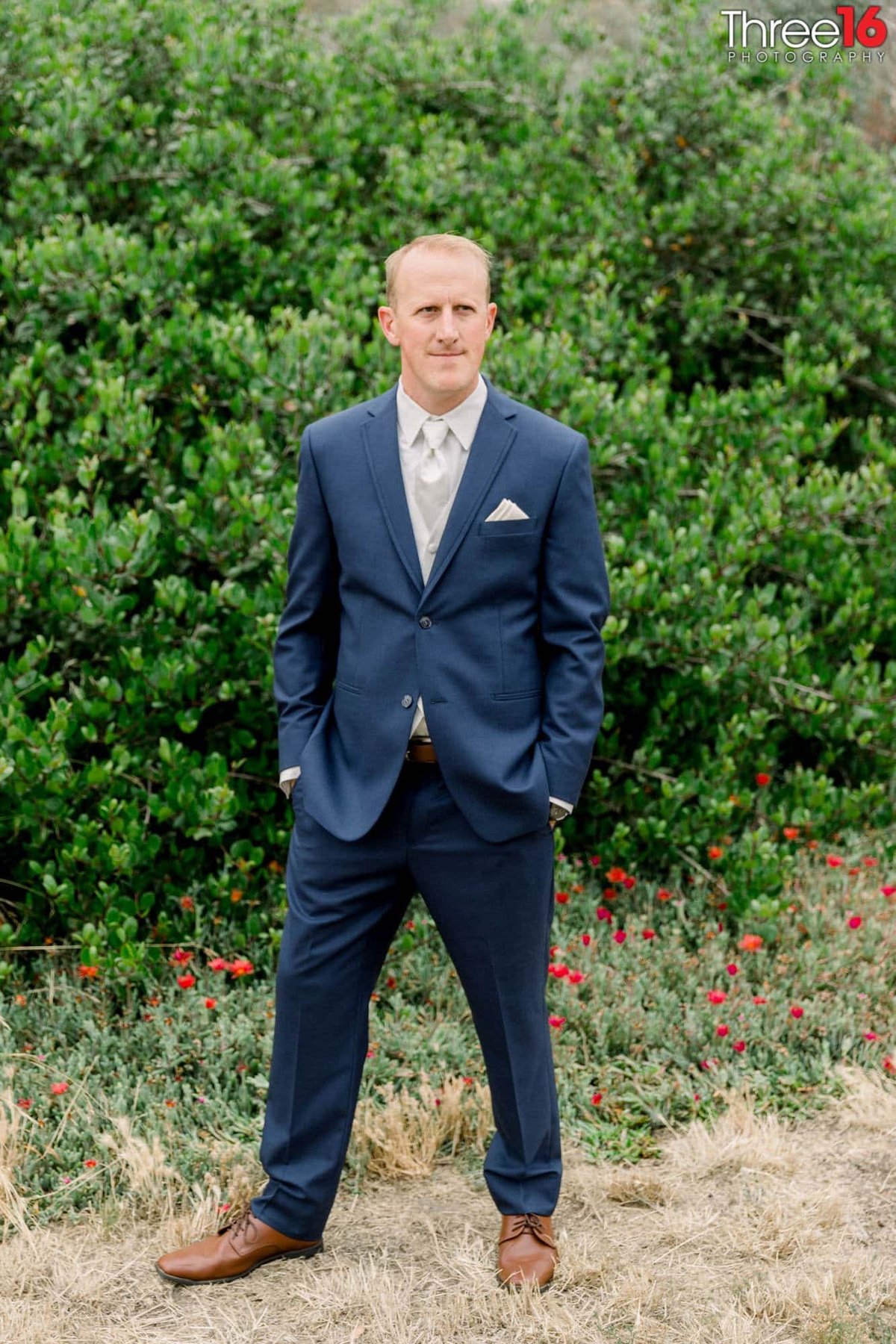 Groom poses for photos  in front of the green shrubs