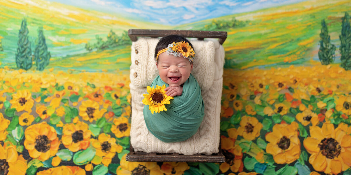 Newborn baby girl wrapped in green and smiling, lying on a tiny bed surrounded by a painted sunflower field backdrop while holding a sunflower.
