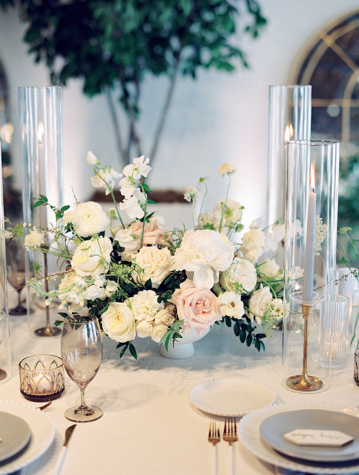 Low floral centerpieces for elegant wedding overflowing with white garden roses, ranunculus, butterfly ranunculus, scabiosa, lisianthus, sweet peas, and natural dark greenery. Floral hues of white, cream, and blush. Designed by Rosemary and Finch in