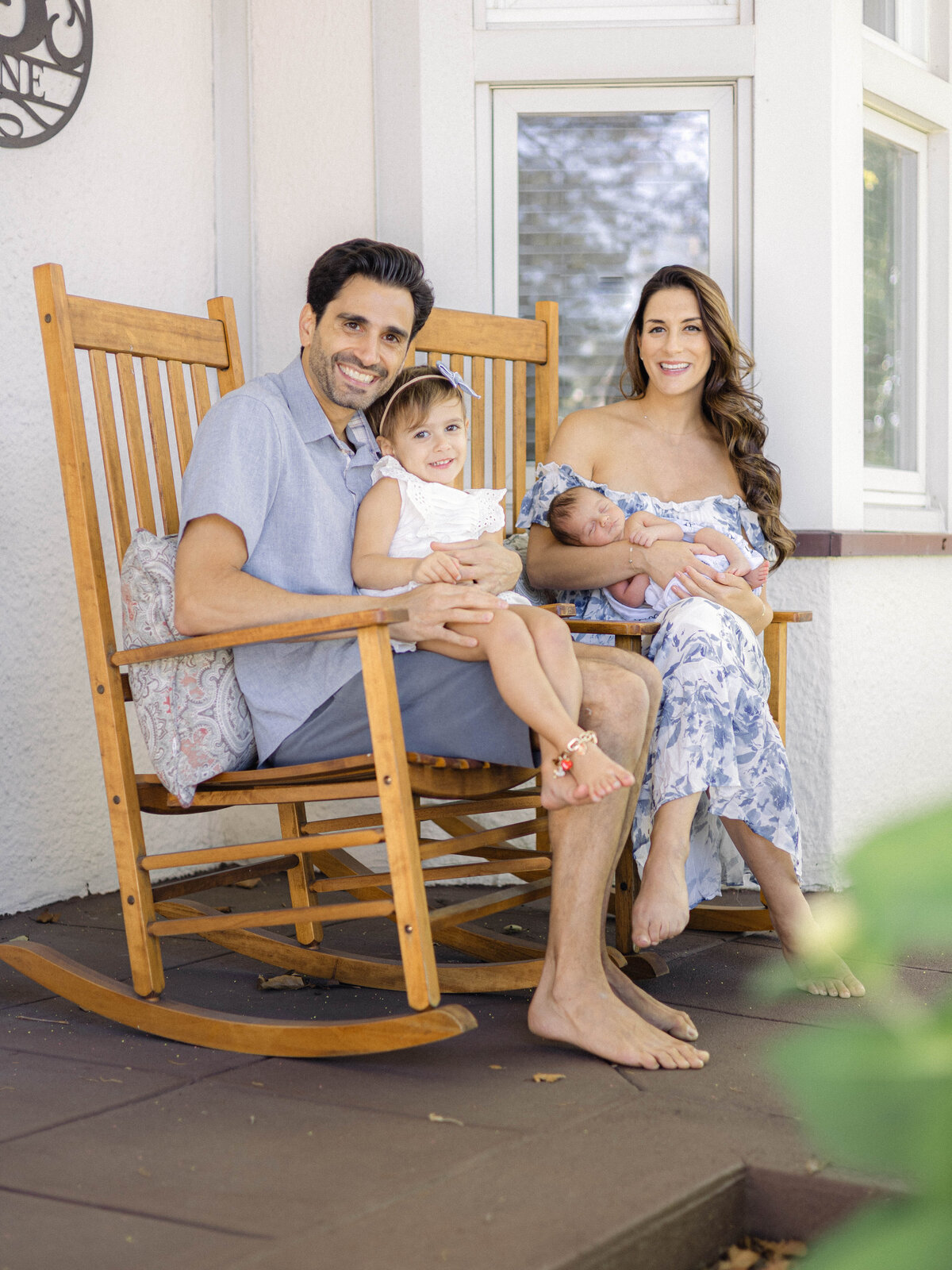 Parents cuddling their newborn and toddler on their front porch.