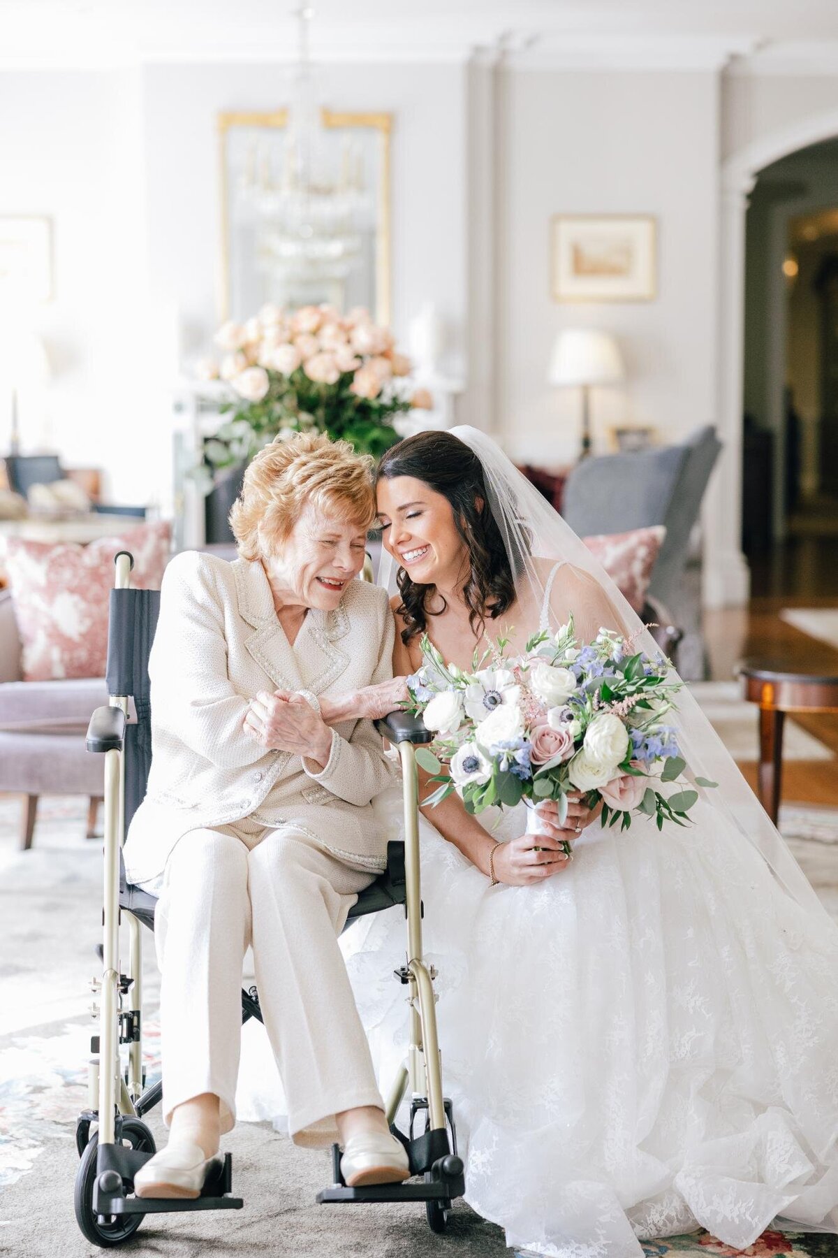 Bride in white dress sharing a tender moment with an elderly woman in a wheelchair, both smiling and holding hands at an indoor setting.