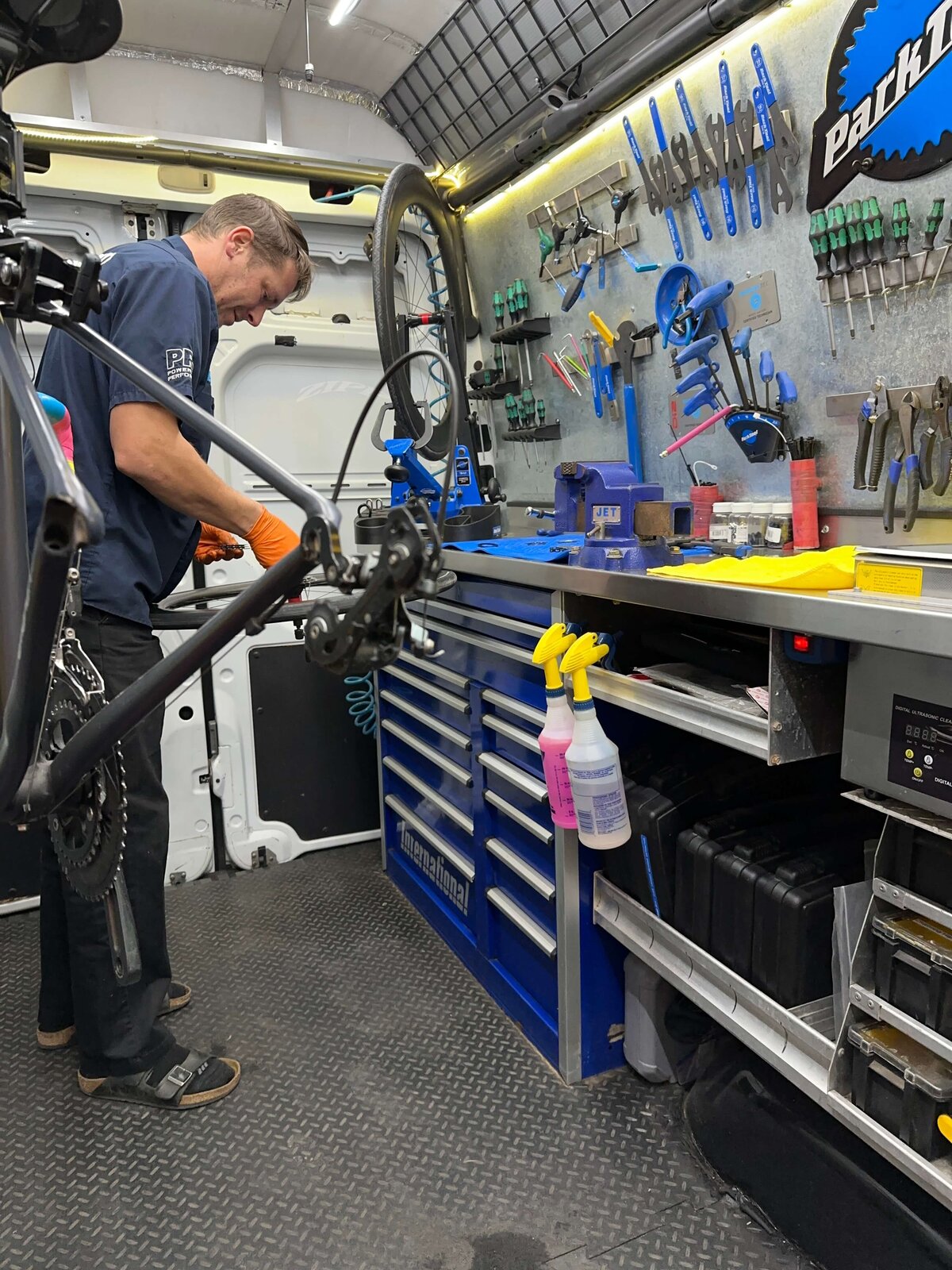 A person in a blue shirt works on a bicycle inside a mobile repair van. Various tools hang on the wall above a blue workbench. Spray bottles are on the edge of the table, and drawers are slightly open below