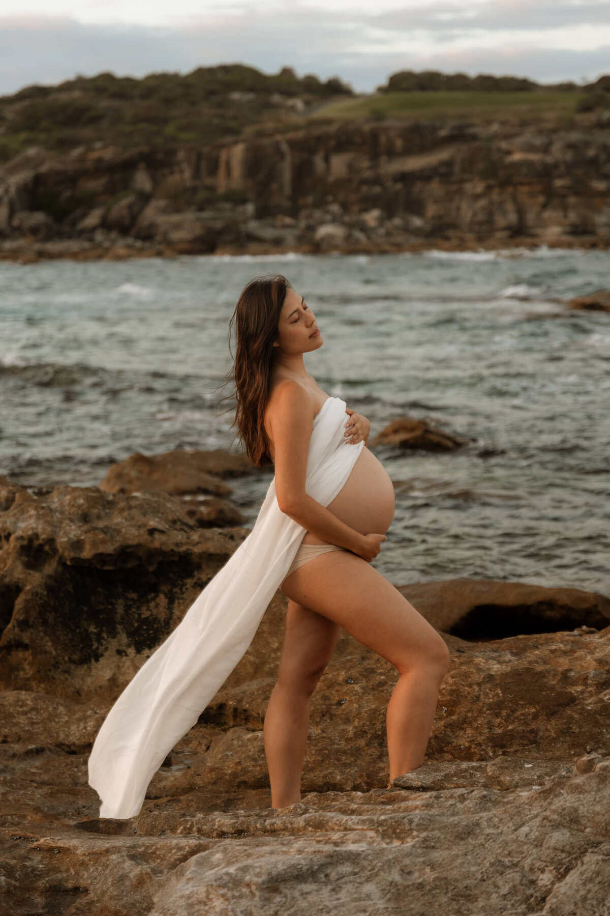 Pregnant woman wrapped in white fabric standing on coastal rocks during a golden hour maternity session in Sydney, Australia.